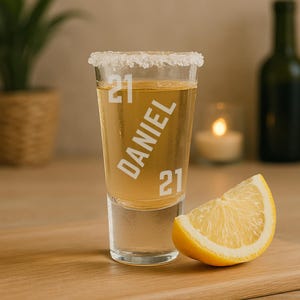 May include: A shot glass with the name "DANIEL" and the number "21" printed on it, filled with a light-coloured liquid and a salted rim. A lemon wedge sits next to the glass. The glass is on a wooden surface.