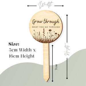 K&ouml;nnte beinhalten: Ein h&ouml;lzerner Gartenpflock mit einem runden Schild, auf dem "Grow through what you go through" steht. Das Schild ist mit einer Silhouette von Wildblumen und Gras verziert. Der Pflock ist 16 cm hoch und 5 cm breit.