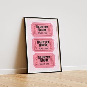 May include: A framed print featuring three pink "Haunted House" tickets. Each ticket has the text "HAUNTED HOUSE ADMIT ONE" and a six-digit number. The tickets are set against a light pink background.
