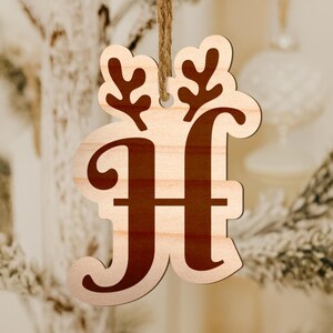 May include: Wooden Christmas ornament shaped like the letter "H" with reindeer antlers. The letter and antlers are a dark brown color, while the background is a light wood. A brown string is attached for hanging.