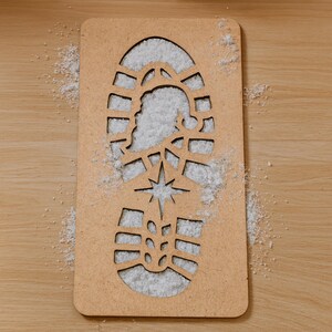 May include: A rectangular, light brown wooden stencil in the shape of a boot print, filled with white material. The stencil features a star and a silhouette of Santa Claus. The background is a light brown wooden surface.