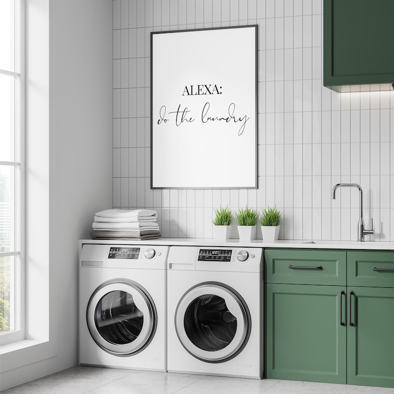 May include: A white framed print with the text "ALEXA: do the laundry" hanging on a white tiled wall above two white washing machines and green cabinets.