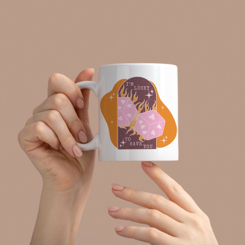 May include: White ceramic mug with a pink and orange design. The design features two dice with hearts on them, flames, and the text "I'm lucky to have you".