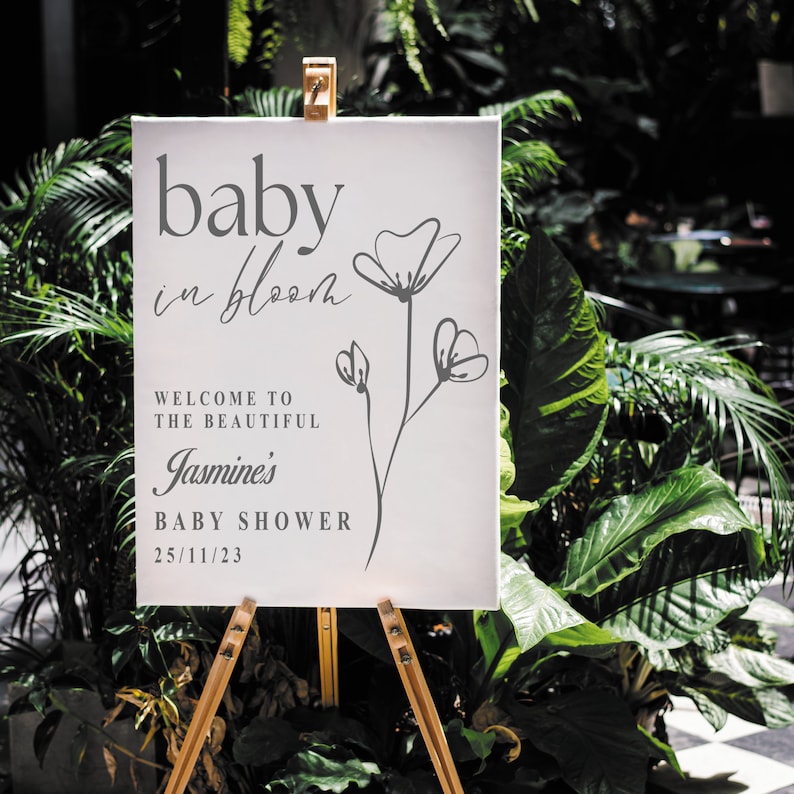 May include: A white sign with the text "baby in bloom" in grey script font. Below the text is a simple line drawing of a flower with three blossoms. The text "Welcome to the beautiful Jasmine's Baby Shower 25/11/23" is also in grey font.