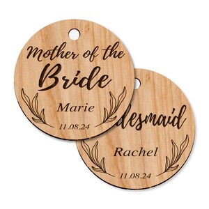 Puede incluir: Dos etiquetas redondas de madera con un dise&ntilde;o floral y el texto "Madre de la novia" y "Dama de honor". La etiqueta para la madre de la novia tiene el nombre "Marie" y la fecha "11.08.24". La etiqueta para la dama de honor tiene el nombre "Rachel" y la fecha "11.08.24".