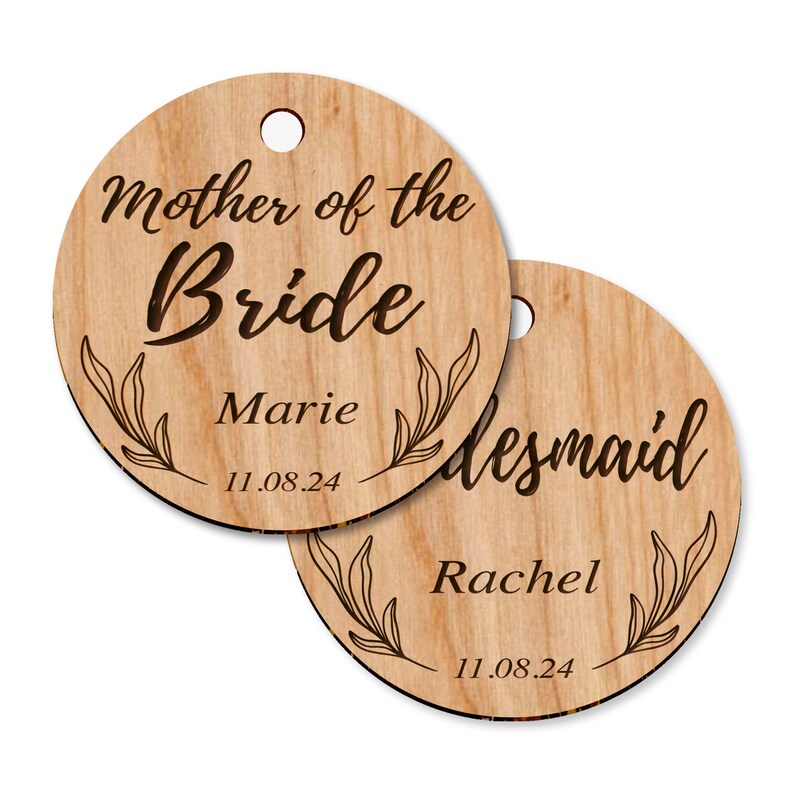 Puede incluir: Dos etiquetas redondas de madera con un dise&ntilde;o floral y el texto "Madre de la novia" y "Dama de honor". La etiqueta para la madre de la novia tiene el nombre "Marie" y la fecha "11.08.24". La etiqueta para la dama de honor tiene el nombre "Rachel" y la fecha "11.08.24".