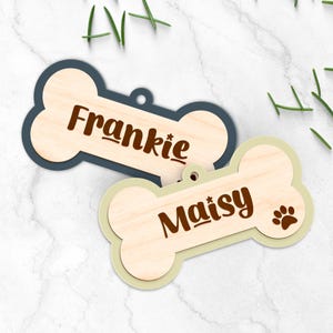 May include: Two bone-shaped pet tags. One tag is navy blue with the name "Frankie" in brown. The other tag is light green with the name "Maisy" and a paw print in brown. Both tags have a light wood grain center.