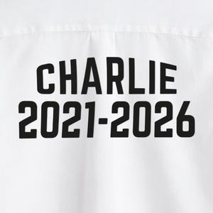Puede incluir: Camiseta blanca con el nombre "CHARLIE" impreso en letras negras en negrita, junto con los a&ntilde;os "2021-2026". El texto est&aacute; centrado y es claramente visible en la prenda.