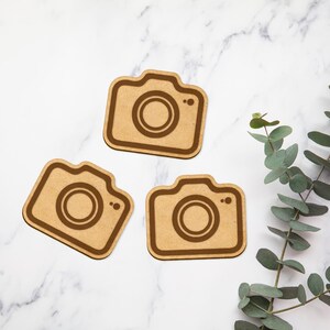 May include: Three wooden camera-shaped ornaments with a brown finish. The ornaments are on a white marble surface.