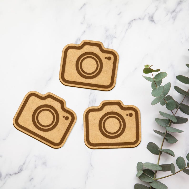 May include: Three wooden camera-shaped ornaments with a brown finish. The ornaments are on a white marble surface.