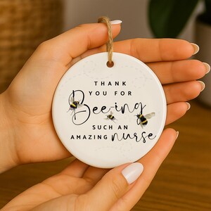 May include: A white ceramic ornament with a brown string, featuring the text "Thank You For Being Such An Amazing Nurse." The design includes illustrations of bees. A thoughtful gift for nurses.