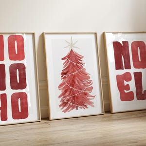 May include: Three framed art prints with a Christmas theme. The prints feature the words "NO HO HO" and "NOEL" in red block letters, and a red watercolor Christmas tree with a star on top. The frames are a light wood color.