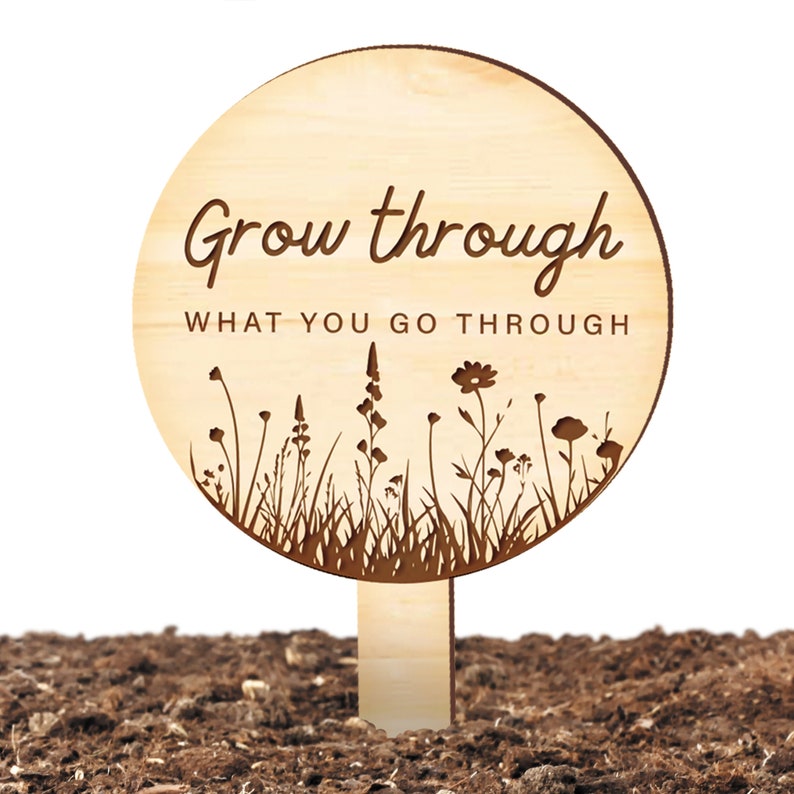 K&ouml;nnte beinhalten: Ein Gartenpfahl aus Holz mit dem Text "Grow through what you go through" und einer Silhouette von Wildblumen.