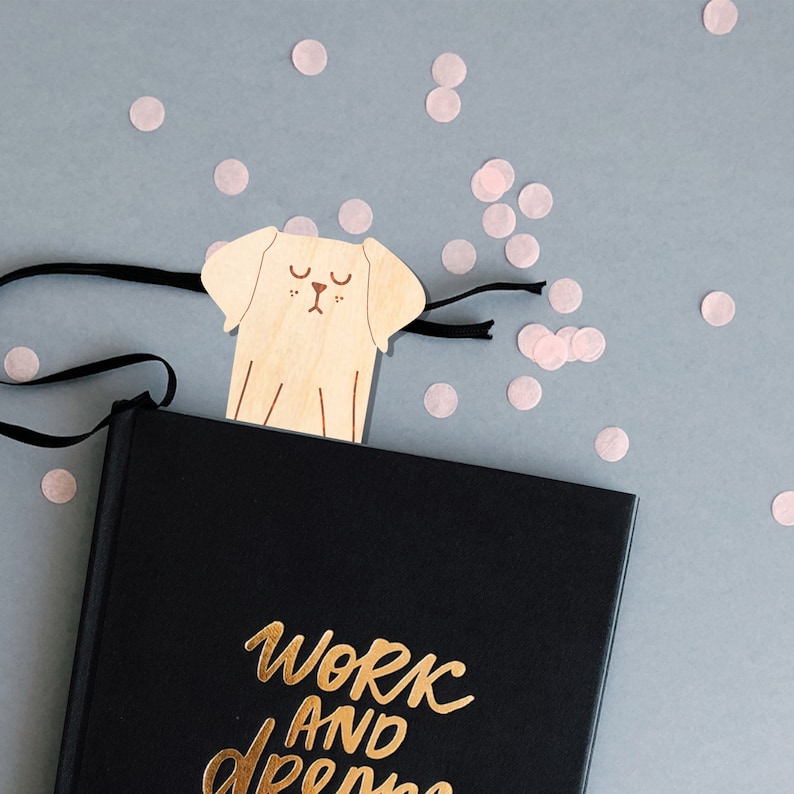 May include: A black journal with the text "Work and Dreams" in gold lettering. A wooden bookmark shaped like a dog is placed on top of the journal.