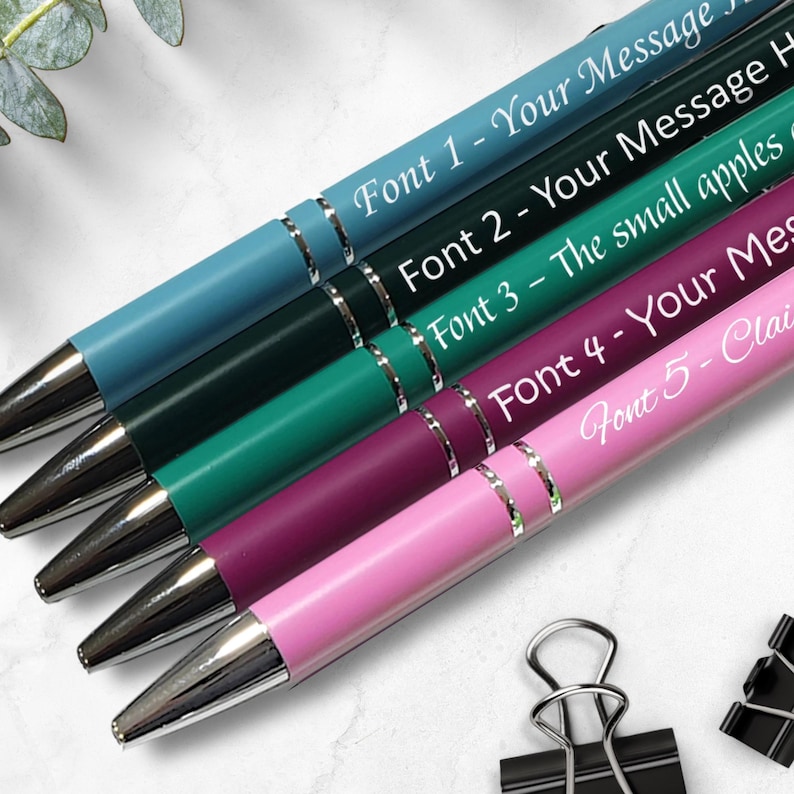 May include: Five pens with different colored barrels and silver accents. Each pen has a different font style and text, including "Font 1 - Your Message", "Font 2 - Your Message", "Font 3 - The small apples", "Font 4 - Your Message", and "Font 5 - Claim".