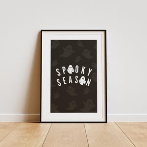 Puede incluir: Una impresi&oacute;n art&iacute;stica enmarcada con un marco negro. La impresi&oacute;n presenta las palabras "Spooky Season" en blanco, con ilustraciones de fantasmas. El fondo es oscuro con un patr&oacute;n de siluetas de fantasmas, ideal para Halloween.