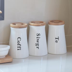 May include: Three white ceramic kitchen canisters with wooden lids. The canisters are labeled "Coffi", "Siwgr", and "Te" in black lettering. The canisters are on a white countertop.