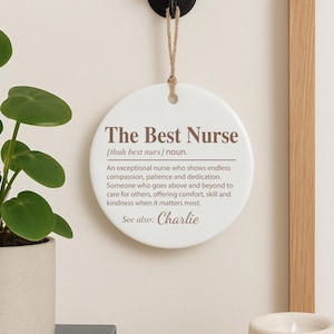 May include: A white, round decorative plaque with the text "The Best Nurse" in brown lettering. The plaque includes a definition of a nurse and the name "Charlie" at the bottom. It hangs from a jute string.