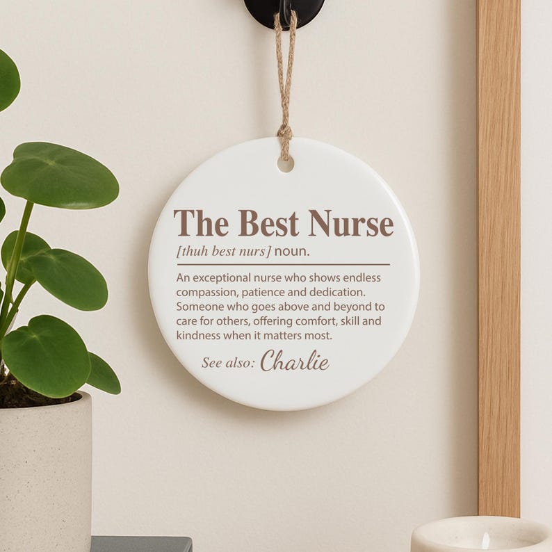 May include: A white, round decorative plaque with the text "The Best Nurse" in brown lettering. The plaque includes a definition of a nurse and the name "Charlie" at the bottom. It hangs from a jute string.