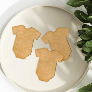 May include: Three wooden baby onesie shapes on a white plate. The onesies are light brown and have a smooth, unfinished surface.