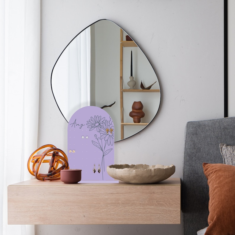 May include: Purple earring holder with a floral design and the name "Amy" written on it. The holder has a rounded top and a flat base. It is sitting on a wooden table with a bowl and a decorative object.