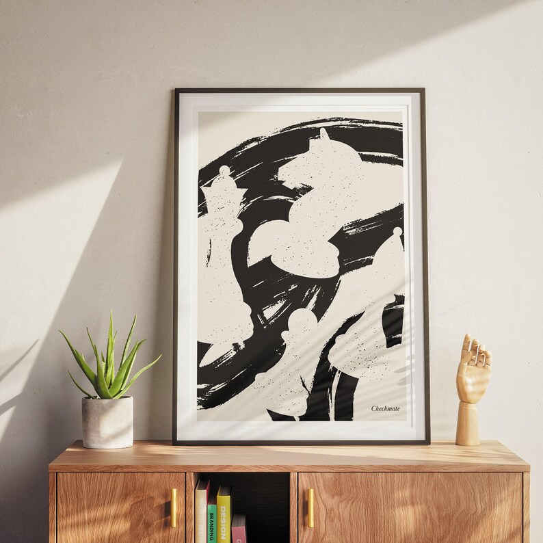 May include: Black and white abstract art print of chess pieces in a minimalist style. The print is framed and hanging on a wall above a wooden shelf with books and a decorative hand.