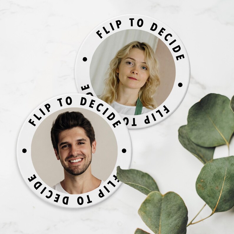 May include: Two white ceramic coasters with the text "FLIP TO DECIDE" encircling a photo of a person. One coaster features a woman, the other a man. Black dots are placed around the edge of the coasters. Eucalyptus leaves are in the background.