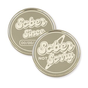 May include: Two silver-colored metal coins with a white design. One coin reads "Sober Since 00/00/" and the other coin reads "Sober Not Sorry".