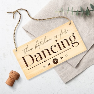May include: Wooden sign with the phrase "This kitchen is for Dancing" in a stylish font. The sign has a music player graphic and is suspended by a natural twine. A cork and a sprig of greenery are also present.