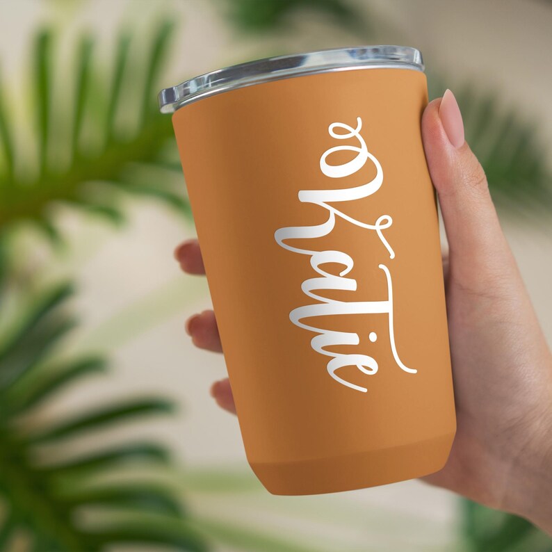 May include: A brown travel mug with a white lid and the word "Katie" written in white cursive on the side.