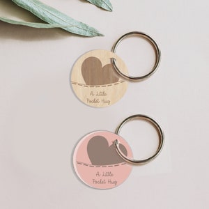 May include: Two round wooden keychains with a brown heart design and the text "A little Pocket Hug". The keychains are attached to silver key rings.