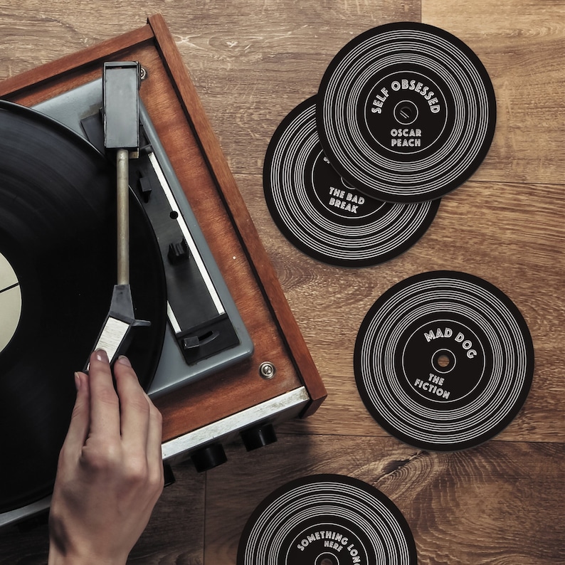 Puede incluir: Cuatro posavasos de vinilo negro y blanco con diferentes nombres de bandas y t&iacute;tulos de &aacute;lbumes. Los posavasos est&aacute;n sobre una superficie de madera junto a un tocadiscos vintage.