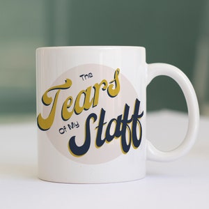 May include: White ceramic mug with a gold and navy blue design. The design features a circle with the text "The Tears of My Staff" in a retro font.