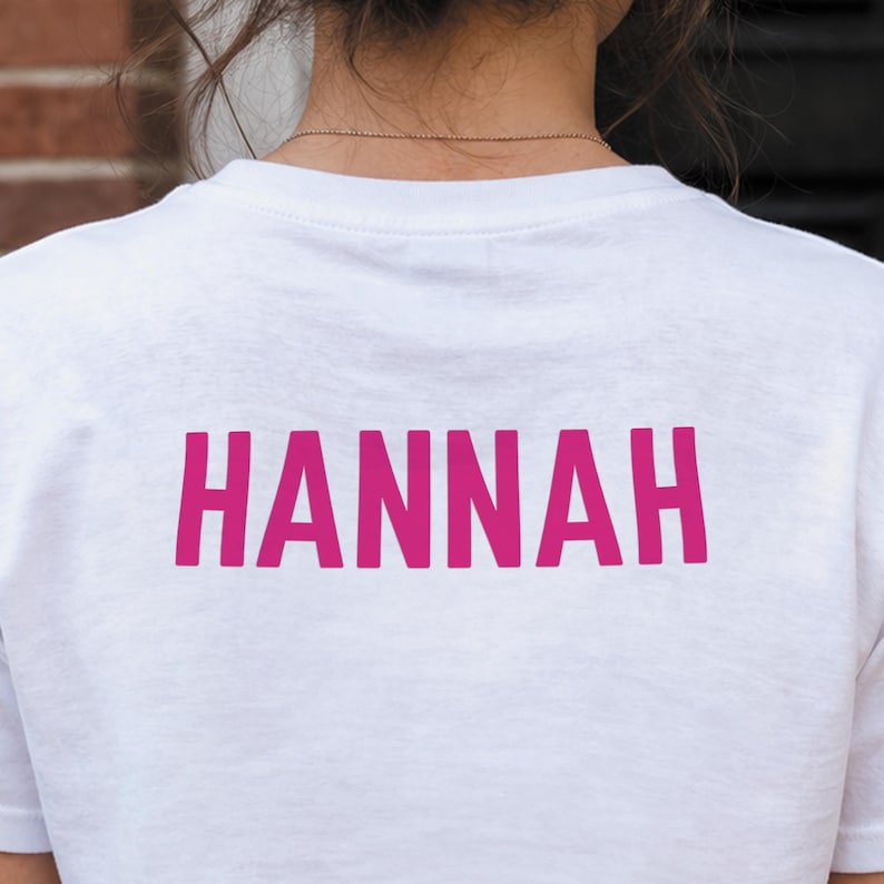 May include: A white t-shirt with the name "HANNAH" printed in a vibrant pink color across the back. The text is in a bold, sans-serif font, making it easily readable. The shirt appears to be made of a soft, comfortable material.