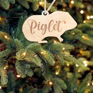 May include: A wooden Christmas ornament shaped like a guinea pig with the name "Piglet" written on it. The ornament is hanging on a green Christmas tree.