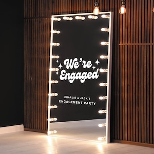 May include: A large, rectangular mirror with a white frame and a black interior. The mirror is illuminated by a series of light bulbs around the perimeter. The text "We're Engaged" is displayed in white script, along with "Charlie & Jack's Engagement Party."