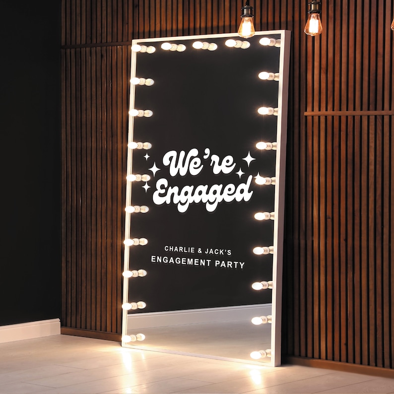 May include: A large, rectangular mirror with a white frame and a black interior. The mirror is illuminated by a series of light bulbs around the perimeter. The text "We're Engaged" is displayed in white script, along with "Charlie & Jack's Engagement Party."