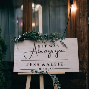 May include: A wooden sign with the text "It was always you" in cursive script, followed by "Jess & Alfie" and the date "08|10|23". The sign is decorated with greenery and is displayed on an easel.