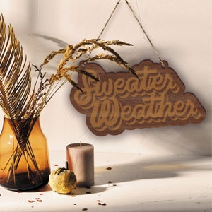 May include: A wooden sign with the words "Sweater Weather" in a layered, cursive font. The sign is brown and hangs from a string. A vase with dried plants, a candle, and a decorative gourd are in the foreground.