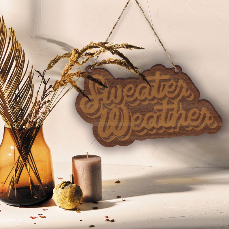 May include: A wooden sign with the words "Sweater Weather" in a layered, cursive font. The sign is brown and hangs from a string. A vase with dried plants, a candle, and a decorative gourd are in the foreground.