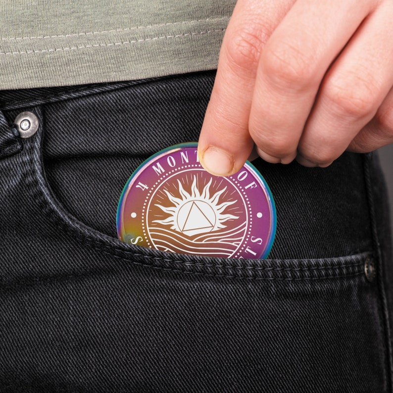 May include: A rainbow iridescent metal coin with the words "The Moment of Silence" and a stylized sun with a triangle inside. The coin is in a pocket of a pair of black jeans.