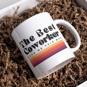 May include: White ceramic mug with a black and white graphic that reads "The Best Coworker Gave Me This Mug" with a retro rainbow stripe design.
