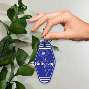 May include: Blue motel key tag with white stripes and a white outline of a flower with the text "Buckle Up Buttercup".