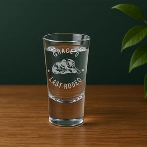 K&ouml;nnte beinhalten: Ein klares Schnapsglas mit Fl&uuml;ssigkeit, mit dem eingravierten Text "Grace's Last Rodeo" und einem Cowboyhut-Design. Das Glas steht auf einer Holzoberfl&auml;che, mit einem dunkelgr&uuml;nen Hintergrund.