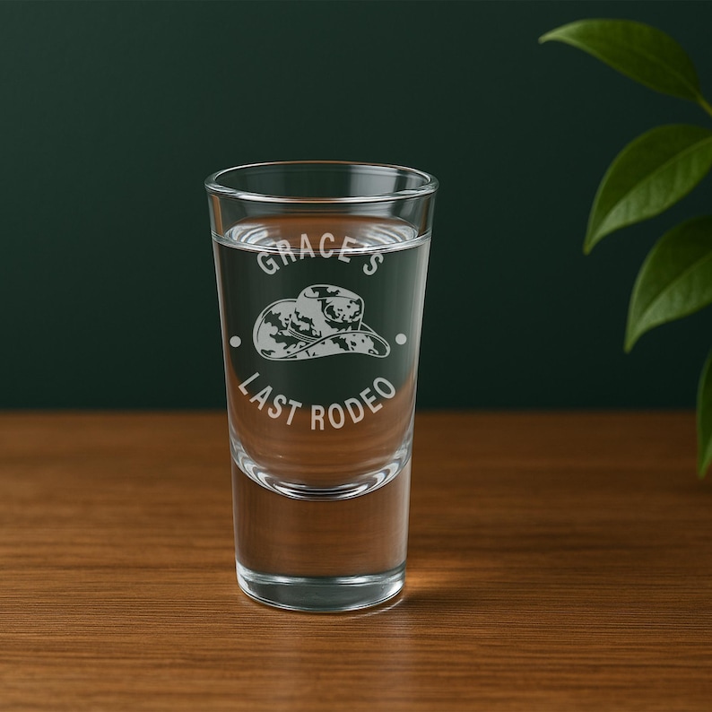 K&ouml;nnte beinhalten: Ein klares Schnapsglas mit Fl&uuml;ssigkeit, mit dem eingravierten Text "Grace's Last Rodeo" und einem Cowboyhut-Design. Das Glas steht auf einer Holzoberfl&auml;che, mit einem dunkelgr&uuml;nen Hintergrund.