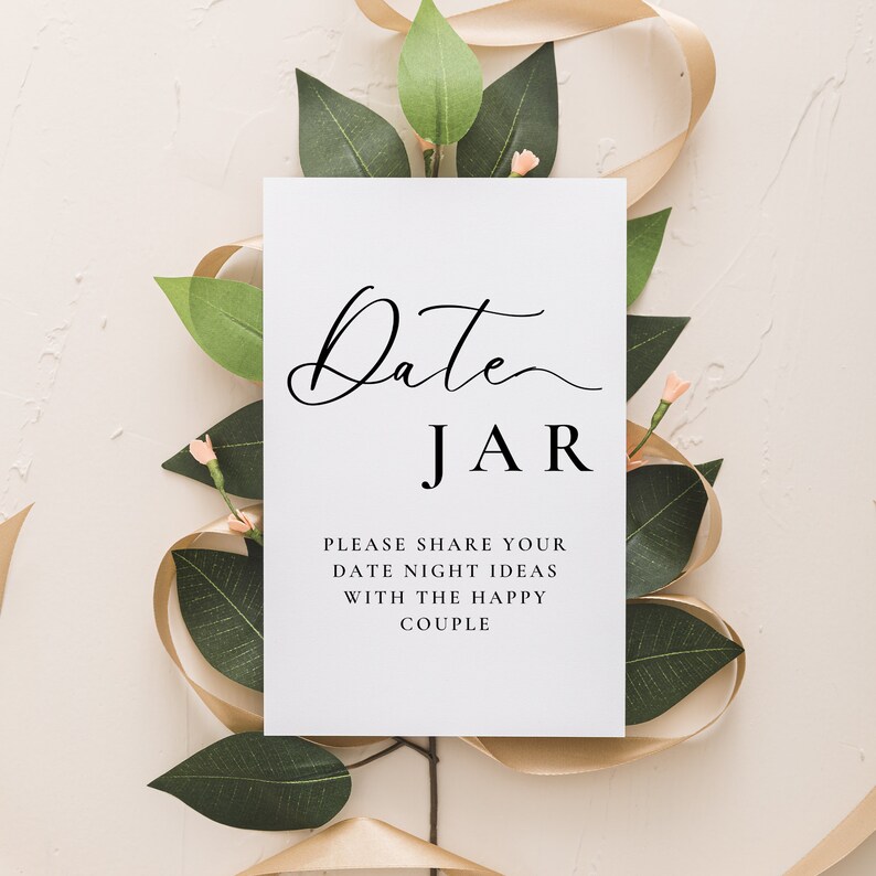 May include: White card with black text that reads "Date JAR" and "Please share your date night ideas with the happy couple". The card is surrounded by green leaves and a gold ribbon.
