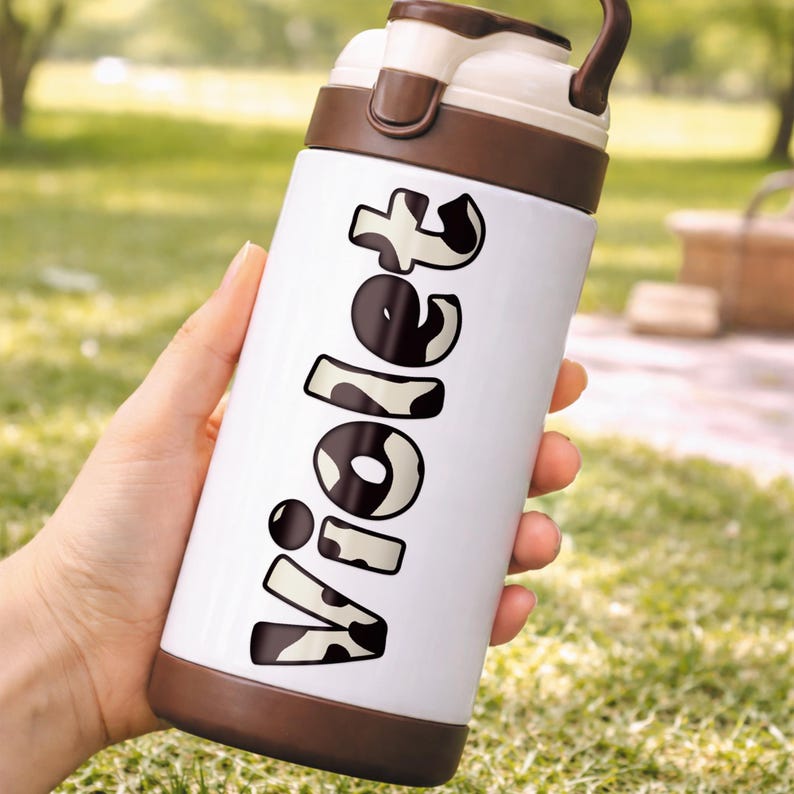 May include: A white insulated water bottle with brown accents and a handle. The word "Violet" is printed vertically in a black and white cow print font. The bottle is held in a person's hand against a blurred green background.