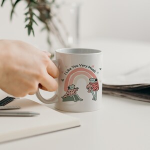 Puede incluir: Taza de cer&aacute;mica blanca con un arco&iacute;ris y dos ranas que llevan jers&eacute;is. El texto en la taza dice "I Like You Very Mush".