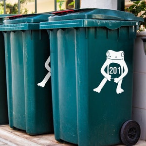 May include: Dark teal wheelie bins with red lids. One bin features a white frog sticker holding a sign with the number 201. The bins are outside, with a pavement and greenery in the background.