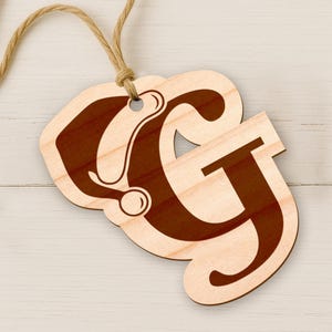 May include: Wooden ornament featuring the letters "CG" in a decorative font, with a dark brown stain on a light wood background. A natural twine loop is attached for hanging. The ornament is on a white wooden surface.